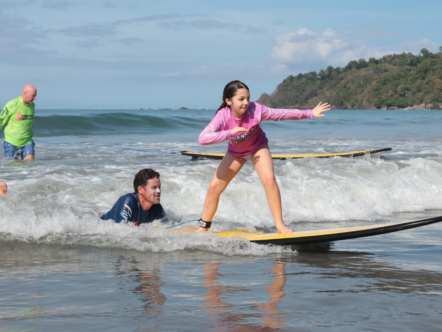 Girl in a pink Dante's rash guard balancing on a yellow surfboard with instructors nearby in Manuel Antonio