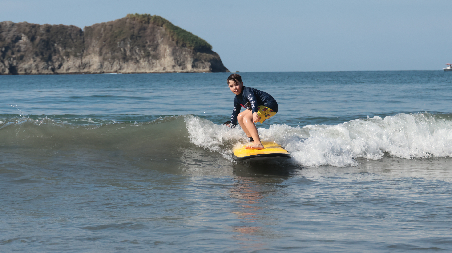Young surfer crouched and riding a wave on a yellow Dante's board with Manuel Antonio's island headland in the background