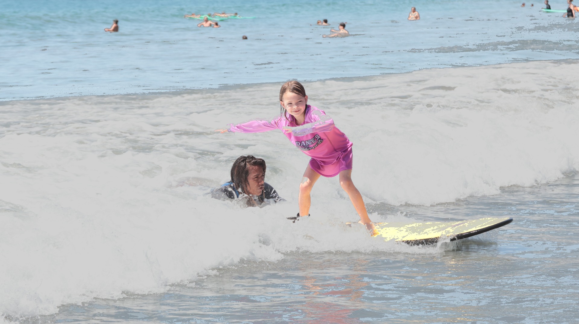 Young girl in a pink Dante's rash guard standing up on a yellow surfboard with her instructor swimming alongside