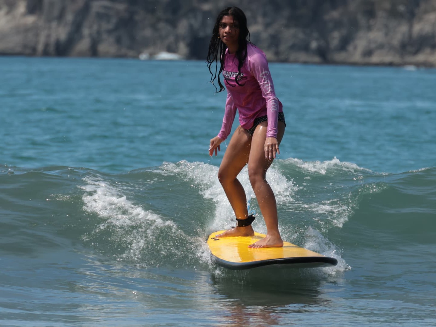 Surfer in a pink Dante's rash guard riding a yellow soft-top with a rocky island headland behind her